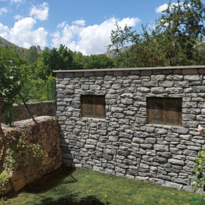 Fachada exterior con revestimiento de piedra gris ceniza instalado en pared, mostrando textura de mampostería natural. Jardín con vegetación y montañas al fondo bajo cielo despejado.