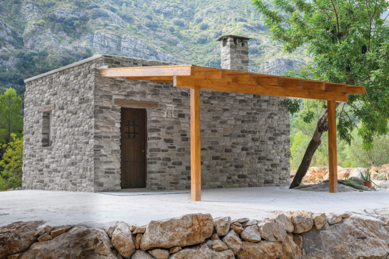 Construcción exterior de revestimiento de piedra gris claro con puerta oscura de madera y porche con estructura de madera, rodeada de vegetación y montañas rocosas al fondo.