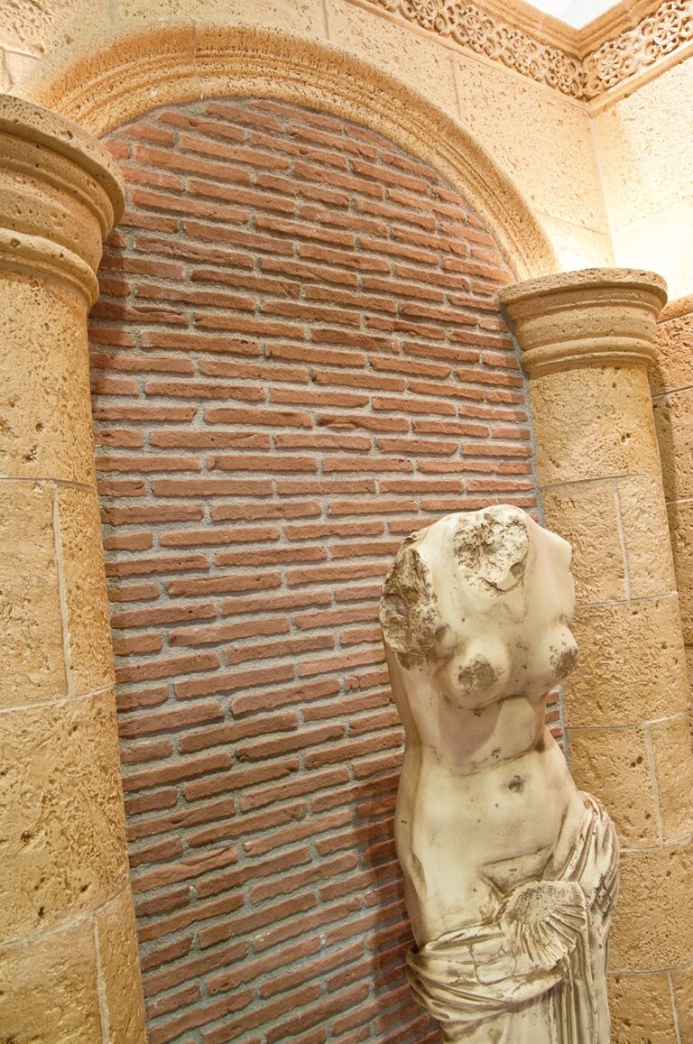 Pasillo interior con arco de piedra y columnas laterales, revestido con ladrillos cerámicos de tono terracota dispuestos horizontalmente. Estatua clásica de torso en mármol blanco junto a la pared derecha, bajo decoración arquitectónica geométrica.