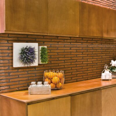 Cocina con revestimiento de pared efecto ladrillo en tonos ocres y marrones, muebles de madera natural, encimera de madera clara y accesorios decorativos con plantas y frutas.