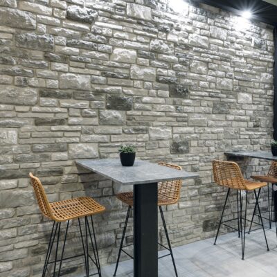 Zona de estar con pared de revestimiento cerámico efecto piedra en tonos grises y ceniza, textura irregular rústica, iluminada con focos LED. Mesa alta de hormigón con base negra y taburetes de ratán con patas metálicas.