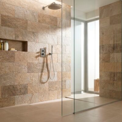 Cuarto de baño contemporáneo con revestimiento de pared de ducha en tonos beige y marrón con textura de piedra natural. Plato de ducha integrado, mampara de cristal sin marco, y nicho empotrado en la pared para accesorios.