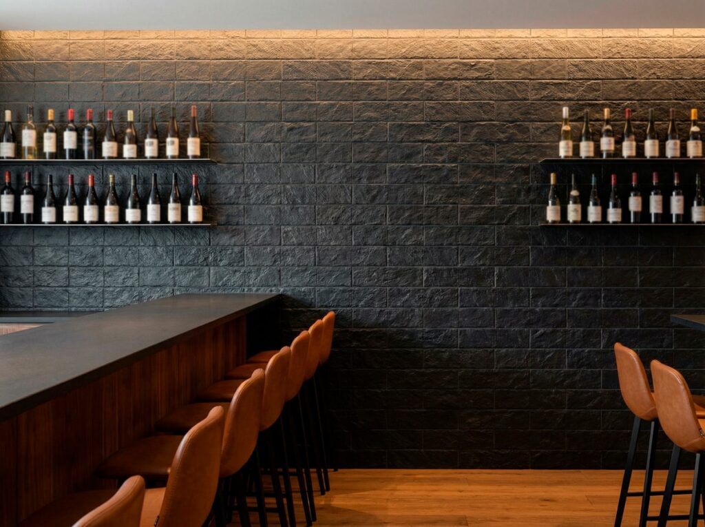 Espacio de bar contemporáneo con revestimiento de pared efecto piedra en tonos gris grafito oscuro, mostradores de madera con asientos tapizados en cuero cognac, y botellas de vino alineadas en estantes iluminados.