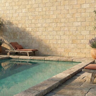Terraza exterior junto a piscina con agua turquesa, revestida con aplacado de hormigón efecto piedra en tonos crema y beige. Mobiliario de descanso y macetas de barro con plantas aromáticas completan el espacio.