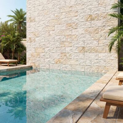 Terraza junto a piscina con pared revestida en piedra artificial Volterra marfil, combinada con suelos porcelanicos, mobiliario de madera y vegetación tropical. Acabado mate con textura irregular que simula piedra natural.