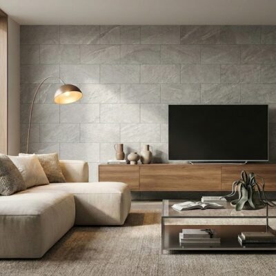Salón contemporáneo con pared de revestimiento gris con textura de piedra. Sofá beige, televisor negro sobre mueble de madera, lámpara de arco dorado y mesa auxiliar de hormigón completan el espacio minimalista.