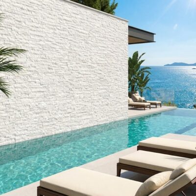 Terraza con piscina infinita frente al mar, revestida con aplacado imitación piedra blanco en textura irregular. Tumbonas beige y plantas verdes complementan el espacio contemporáneo junto a la costa.