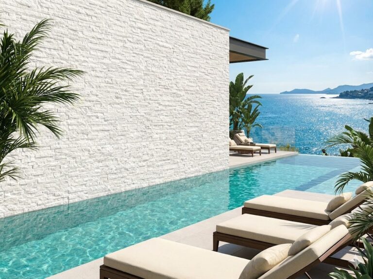 Terraza con piscina infinita frente al mar, revestida con aplacado imitación piedra blanco en textura irregular. Tumbonas beige y plantas verdes complementan el espacio contemporáneo junto a la costa.