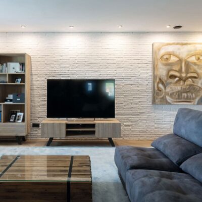 Salón contemporáneo con pared de revestimiento imitación ladrillo blanco texturizado, televisor empotrado en estantería de madera, sofá azul oscuro y arte mural dorado.