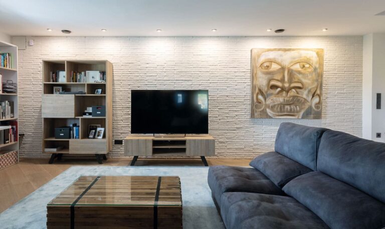 Salón contemporáneo con pared de revestimiento imitación ladrillo blanco texturizado, televisor empotrado en estantería de madera, sofá azul oscuro y arte mural dorado.