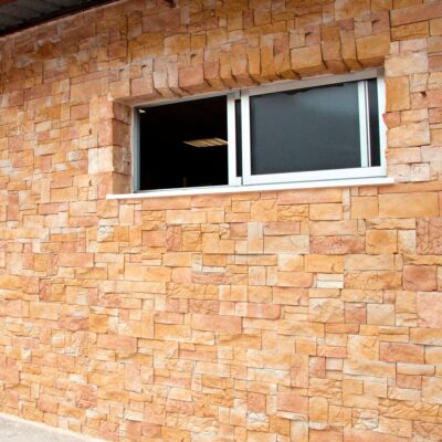 Fachada de edificio revestida con aplacado imitación piedra en tonos beige y crema, con textura irregular y relieve. Ventana horizontal con marco metálico plateado integrada en el revestimiento.