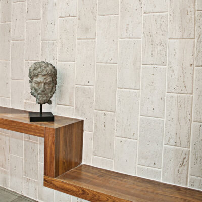 Nicho de pared con revestimiento imitación piedra blanca travertino en formato rectangular, instalado en herringbone. Pedestal de madera oscura muestra busto de escayola gris contra el muro texturizado.