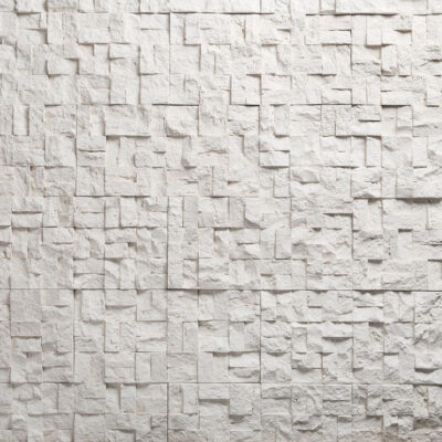 Superficie con textura de piedra blanca en relieve, formada por piezas irregulares de diferentes tamaños que crean profundidad y sombras. Acabado mate con tonos beige y gris claro.