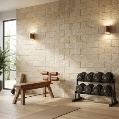 Espacio de entrenamiento con revestimiento de pared imitación piedra en tonos beige y crema. Visible banco de pesas de madera, mancuernas en soporte metálico, saco de boxeo rojo y equipamiento deportivo sobre suelo de gres porcelánico claro.