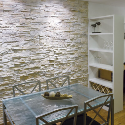 Salón contemporáneo con pared de revestimiento imitación piedra en tonos blancos y beige, acabado texturizado. Mobiliario moderno con mesa de cristal y sillas de madera, complementado con estanterías blancas de almacenamiento.