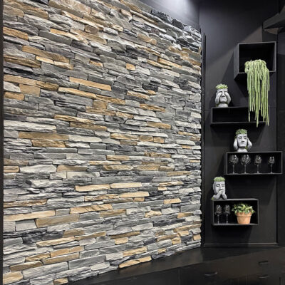 Pared de salón revestida con baldosas imitación piedra natural en tonos grises y beige, instaladas en formato rectangular. A la derecha, estantería negra con plantas decorativas y accesorios, iluminada por focos LED. Mueblería negra inferior completa el espacio.