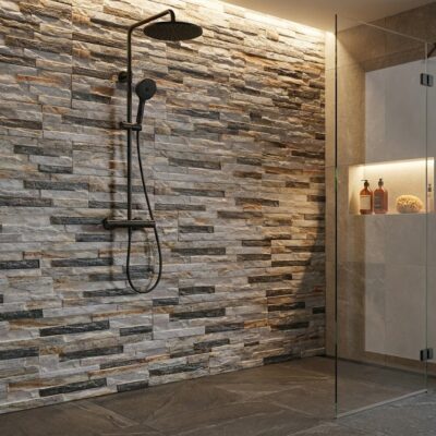 Ducha interior con revestimiento de pared imitación piedra en tonos grises y beige, acabado natural mate. Panel de cristal transparente, grifo de ducha negro matte, nicho iluminado con productos de baño y decoración.