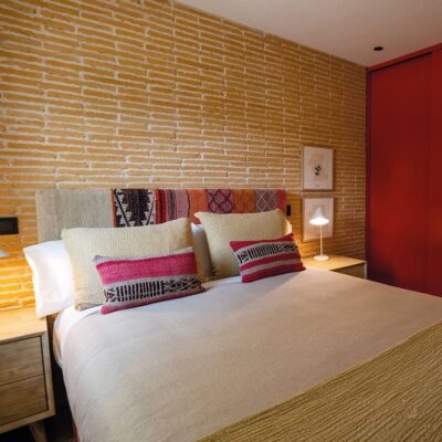 Dormitorio con pared de revestimiento interior imitación ladrillo en tono rojizo cálido, complementado con detalles en rojo intenso y textiles de colores tierra. La pared de ladrillo cubre el cabecero y proporciona textura visual al espacio.