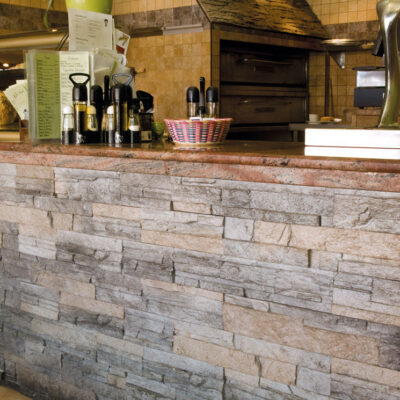 Mostrador de comercio revestido con aplacado imitación piedra en tonos grises y beige, con textura irregular apilada. Encimera de granito, equipamiento de negocio visible al fondo y azulejos de pared en tonos cálidos.