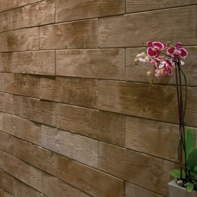Revestimiento de pared Landes Verniprens con acabado imitación madera en tonos beige y marrones, acompañado de una orquídea fucsia en maceta blanca. El material presenta textura de veta de madera natural y formato de piezas rectangulares horizontales.
