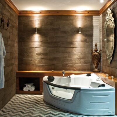 Cuarto de baño elegante revestido con baldosas porcelánicas imitación madera en tonos oscuros. La pared posterior presenta formato rectangular grande con textura de madera envejecida, iluminada con apliques empotrados. Suelo con patrón geométrico en blanco y gris, bañera angular blanca, espejo ovalado dorado y detalles decorativos en bronce.