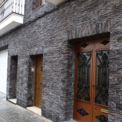 Fachada exterior con aplacado de revestimiento gris oscuro tipo piedra natural. La pared muestra textura de mamposterÍa con piezas irregulares, complementada por puertas de madera y herrajes decorativos de forja en la entrada principal.