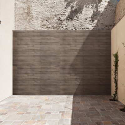 Terraza exterior con revestimiento de pared en listón Montreal de tonalidad gris oscura, suelo de gres en tonos beige y terracota, plantas trepadoras en maceta y acceso a vivienda mediante puerta-ventana oscura.