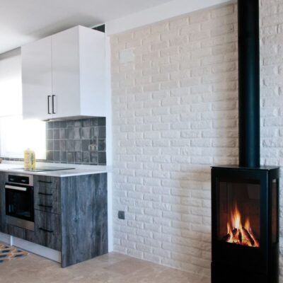 Cocina contemporánea con revestimiento de pared London blanco de textura ladrillo, muebles inferiores en tonos azulados y muebles superiores blancos. Estufa de leña negra integrada en la pared lateral derecha.
