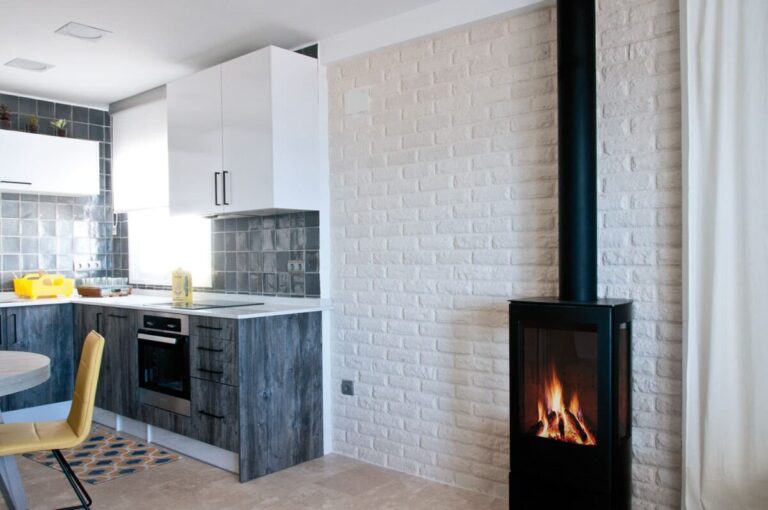 Cocina contemporánea con revestimiento de pared London blanco de textura ladrillo, muebles inferiores en tonos azulados y muebles superiores blancos. Estufa de leña negra integrada en la pared lateral derecha.