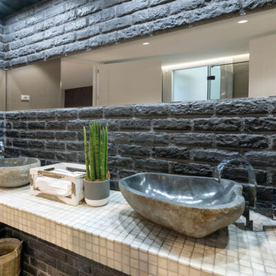 Baño contemporáneo con paredes revestidas de piedra negra texturizada y suelo de mosaico blanco. Dos lavabos de piedra natural sobre encimera blanca, grifo negro y espejo grande. Techo con vigas de hormigón expuesto.