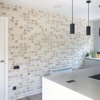 Cocina contemporánea con pared de revestimiento ladrillo blanco tipo London Polar, isla central blanca, pendientes negros cilíndricos y ventana con vistas al exterior.