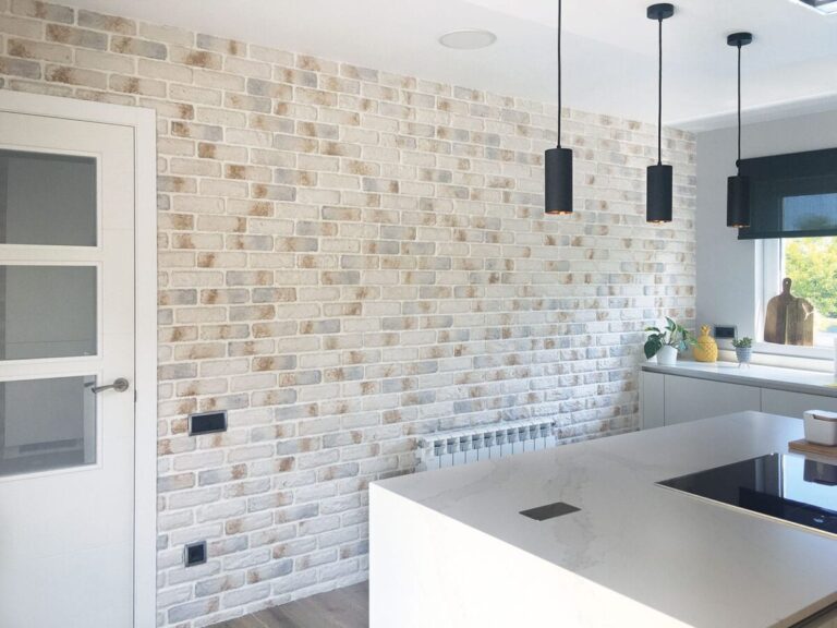 Cocina contemporánea con pared de revestimiento ladrillo blanco tipo London Polar, isla central blanca, pendientes negros cilíndricos y ventana con vistas al exterior.