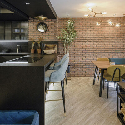 Cocina contemporánea con paredes de ladrillo visto en tono rojizo y revestimiento London Támesis. Isla negra con encimera gris, taburetes tapizados en gris y verde, mesa de madera con sillas verdes, plantas decorativas y luminaria dorada de diseño moderno.