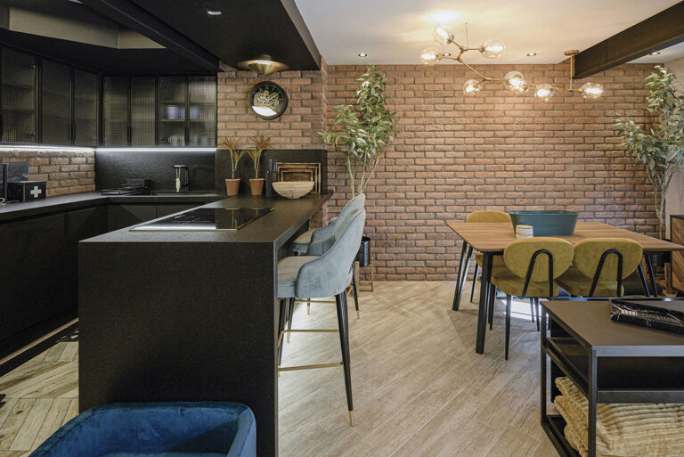 Cocina contemporánea con paredes de ladrillo visto en tono rojizo y revestimiento London Támesis. Isla negra con encimera gris, taburetes tapizados en gris y verde, mesa de madera con sillas verdes, plantas decorativas y luminaria dorada de diseño moderno.