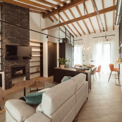 Salón diáfano con suelo de gres en tonos claros, revestimiento de pared tipo ladrillo en la chimenea central, vigas de madera vista en el techo y zona de comedor integrada con sillas tapizadas en naranja.