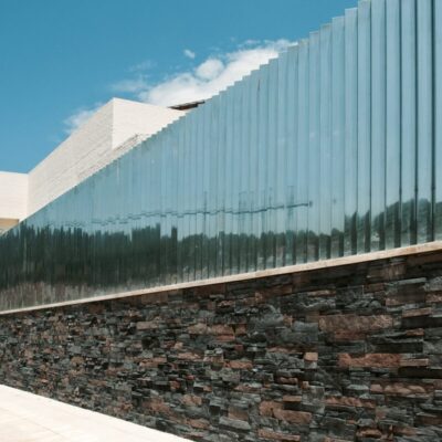 Fachada exterior contemporánea con aplacado de piedra natural en tonos grises y marrones en la base, combinado con paneles de revestimiento gris claro en la parte superior. La composición muestra una arquitectura minimalista con líneas horizontales pronunciadas bajo cielo despejado.