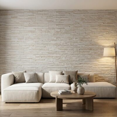 Salón contemporáneo con pared de revestimiento moderno efecto piedra en tonos crema y beige. Sofá modular de lino claro, mesa de centro ovalada en madera natural, suelo de gres en formato madera clara, y plantas decorativas complementan el espacio luminoso.