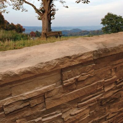 Detalle de muro exterior con aplacado imitación piedra en tonos cálidos beige y marrón, instalado en un espacio abierto de jardín con vistas panorámicas al valle y montañas.