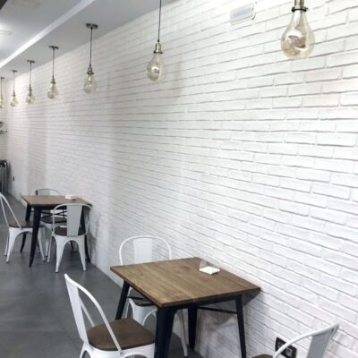 Espacio de comedor contemporáneo con paredes revestidas en azulejos blancos con textura de ladrillo. Mesas de madera con patas metálicas y sillas blancas, iluminación con lámparas colgantes de vidrio ámbar.