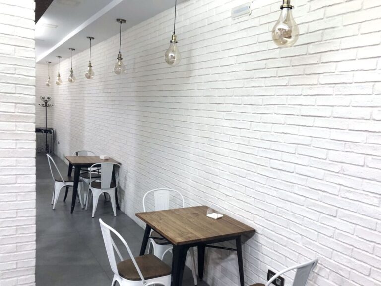 Espacio de comedor contemporáneo con paredes revestidas en azulejos blancos con textura de ladrillo. Mesas de madera con patas metálicas y sillas blancas, iluminación con lámparas colgantes de vidrio ámbar.