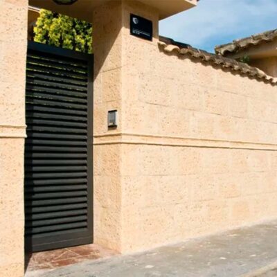 Fachada exterior con revestimiento para muros color beige envejecido, textura rústica tipo piedra, con puerta metálica negra y portón de garaje, bajo luz natural diurna.