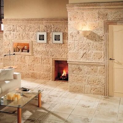 Salón con paredes revestidas con imitación piedra tosca en tonos beige y crema, chimenea integrada con fuego encendido, suelo cerámico claro y puerta de entrada crema.