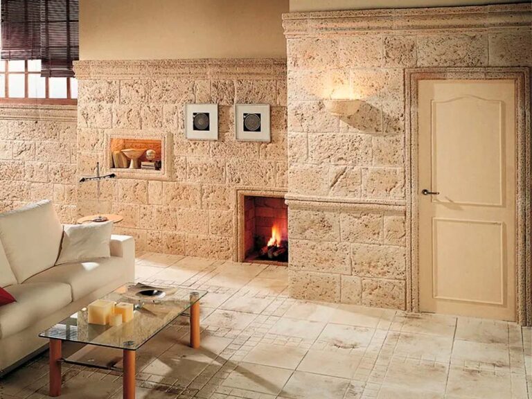 Salón con paredes revestidas con imitación piedra tosca en tonos beige y crema, chimenea integrada con fuego encendido, suelo cerámico claro y puerta de entrada crema.