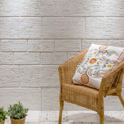 Pared interior con revestimiento en tonos marfil que imita piedra natural, combinada con una silla de mimbre y macetas decorativas en primer plano.