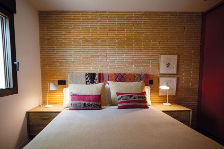 Dormitorio con pared de revestimiento que imita ladrillo caravista en tonos cálidos Manchester Amanecer, cama con almohadas decorativas en rojo y crema, y lámparas de lectura a ambos lados del cabecero.