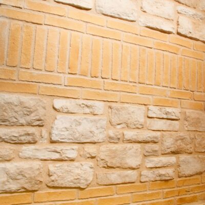 Dos muestras de revestimiento de pared: superior con efecto ladrillo cerámico fino en tonos dorados y beige, inferior con textura de piedra natural en tonos crema y arena. Ambas presentan acabado rústico con relieve.