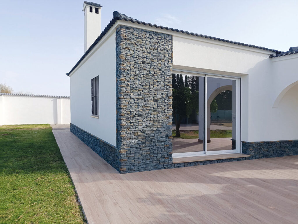 Vivienda moderna con fachada lateral revestida en aplacado de hormigón imitación piedra gris marengo, gran formato rectangular. Junto a pared blanca y chimenea de hormigón, con terraza de suelo porcelánico y jardín.