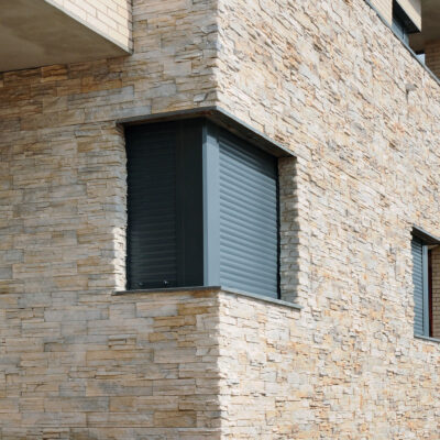Fachada de edificio revestida con aplacado que imita piedra en tonos beige y gris, con persianas grises en ventanas. El revestimiento exterior muestra acabado rugoso y texturizado característico de piedra natural.