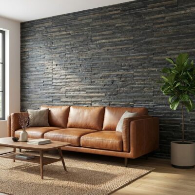 Salón contemporáneo con sofá de cuero marrón y pared de revestimiento efecto piedra gris grafito en formato 10x50 cm. Junto a la ventana, planta de interior en maceta complementa el espacio minimalista con suelo de madera y alfombra textil.