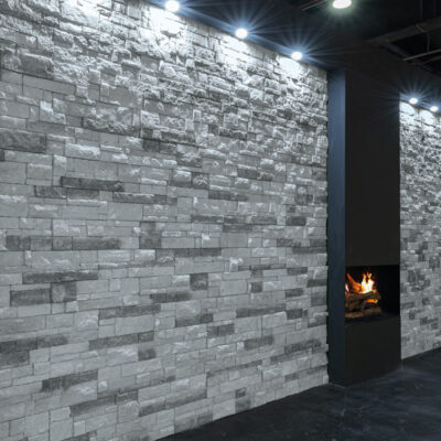 Pared interior revestida con aplacado de piedra gris en tonos ceniza y blanco, con formato rectangular en patrón de soga. Luminarias empotradas iluminan la textura rugosa. Chimenea moderna integrada en el lateral derecho con fuego visible.